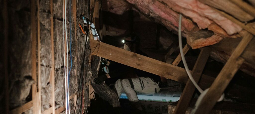 A certified insulation installer works in a crammed roof space, surrounded by timber joists and electrical wires.