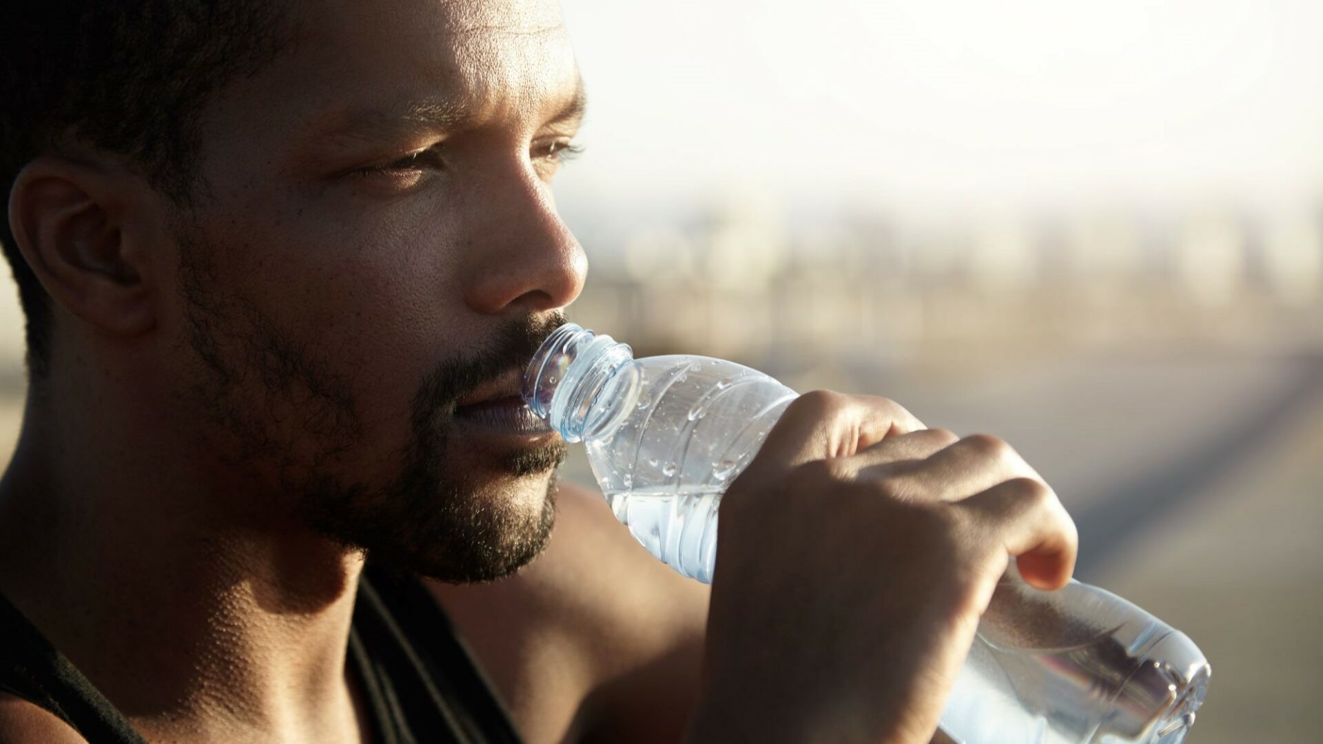 A dark-skinned, male tradie looks into the distance while drinking water.